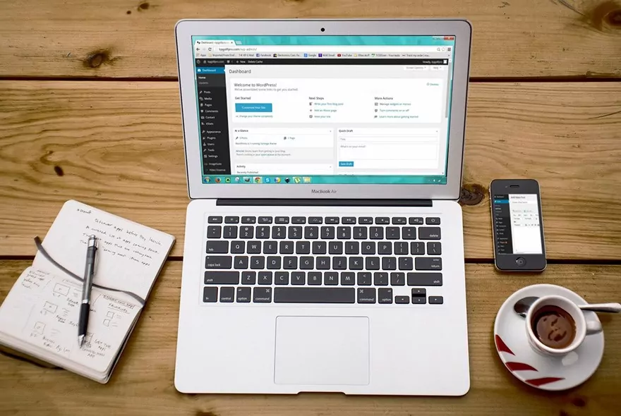 Laptop displaying WordPress dashboard alongside a notepad, pen, smartphone, and coffee cup on a wooden table, illustrating digital marketing and web design workflow.